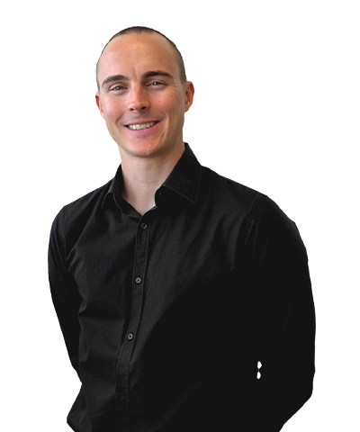 Adam Barbarich, with short hair and a black button-up shirt, stands against a plain, light background, smiling with hands behind their back.