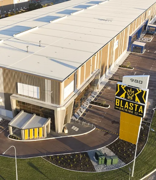 Aerial view of a large modern building, known as the SARICH Building, with a tall sign reading Blasta Brewing Co. at the entrance. The facility features a white roof, tan exterior, and landscaped grounds.