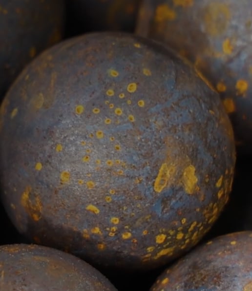 A close-up view of a round, dark surface with scattered yellow and orange patches, resembling rust or oxidation spots on grinding media used in the mining industry. Other similar spheres are partially visible in the background.