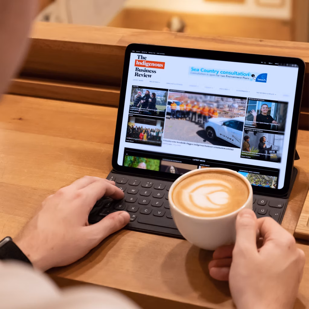 Person holding a cup of coffee and viewing Indigenous news on The Indigenous Business Review website, using a tablet with a keyboard case while seated at a wooden table.