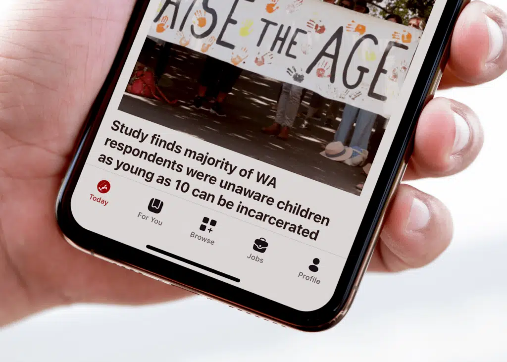 A hand holds a mobile phone displaying an Aboriginal news story from the National Indigenous Times about a study on WA respondents’ awareness of child imprisonment. The screen shows part of a banner reading RAISE THE AGE with colourful handprints.