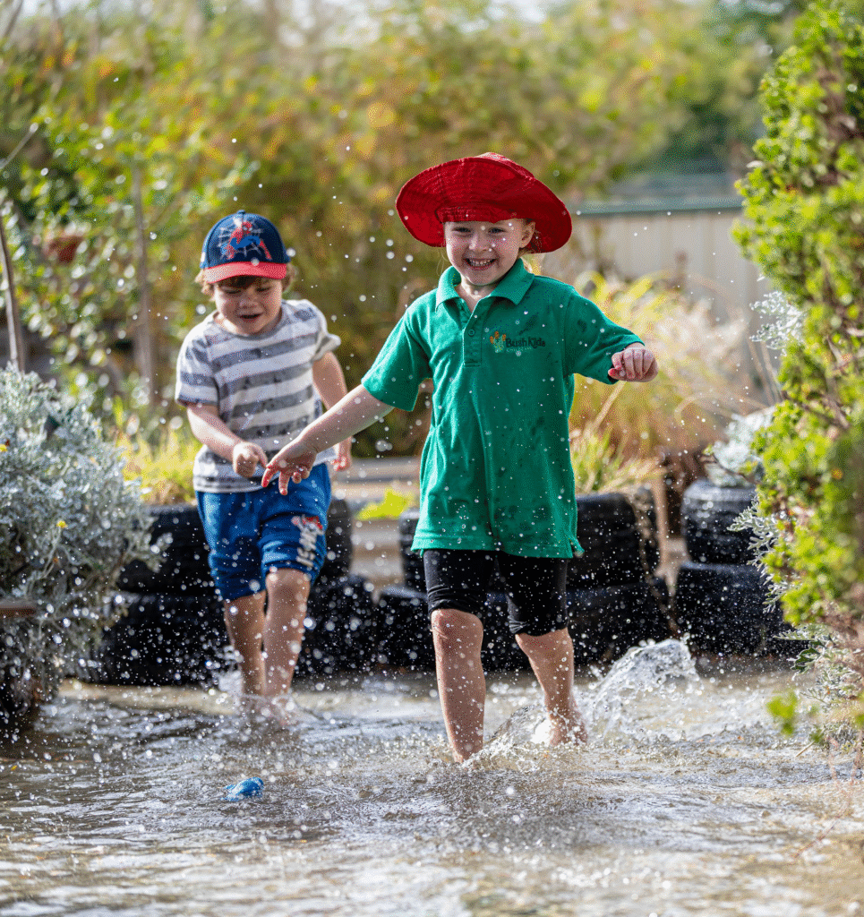 Two young children, one in a red hat and green shirt, the other in a blue cap and striped shirt, joyfully splash and run through shallow water outdoors, surrounded by greenery and sunlight.