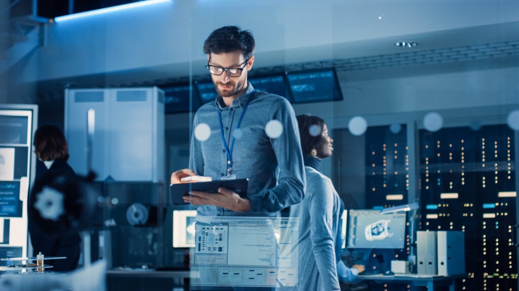 A man wearing glasses stands in a modern office, looking at a tablet. Computer screens, servers, and a woman working in the background suggest a high-tech or cyber-security environment in the technology sector.