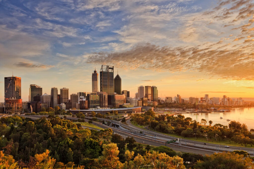 A city skyline at sunset with tall buildings, a river, and motorways surrounded by greenery, under a partly cloudy sky with warm sunlight illuminating the scene.