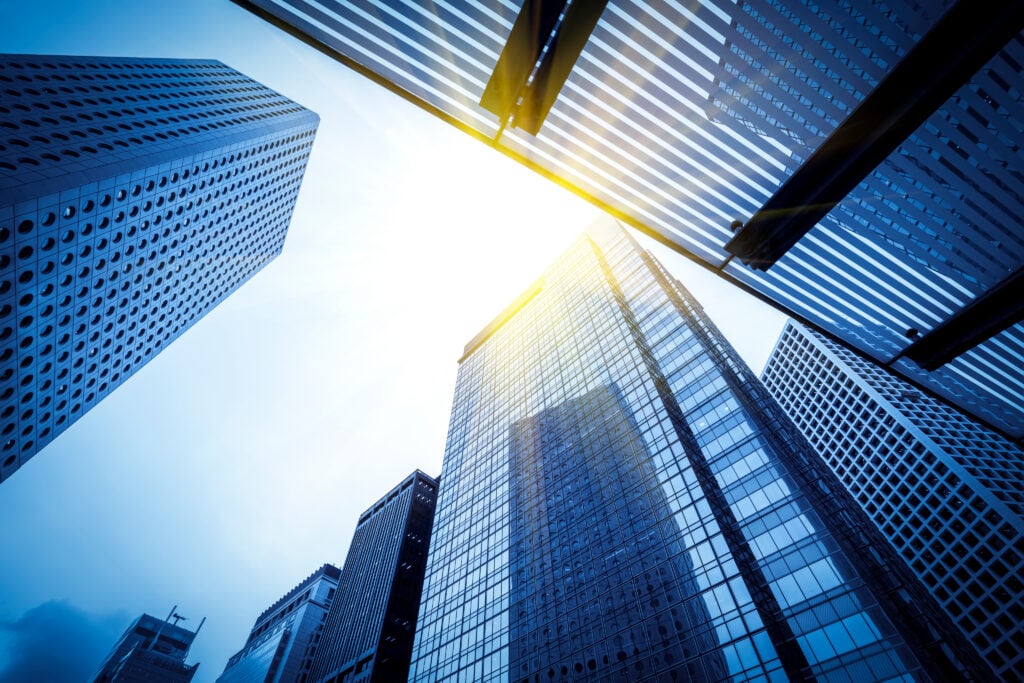 Skyscrapers with glass and steel façades rise towards a bright sky, reflecting sunlight. The photo is taken from a low angle, emphasising the height and modern architecture of the buildings.