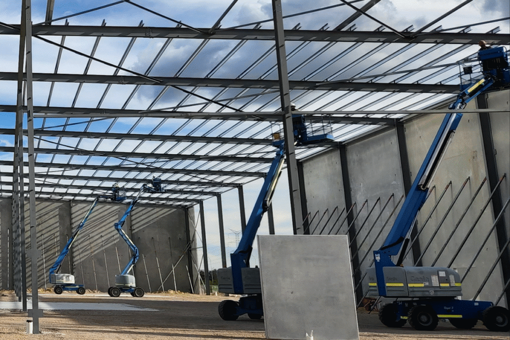 Several blue cherry pickers raise workers during construction to install metal beams on the roof of a large industrial steel structure, with concrete walls and a cloudy sky in the background.