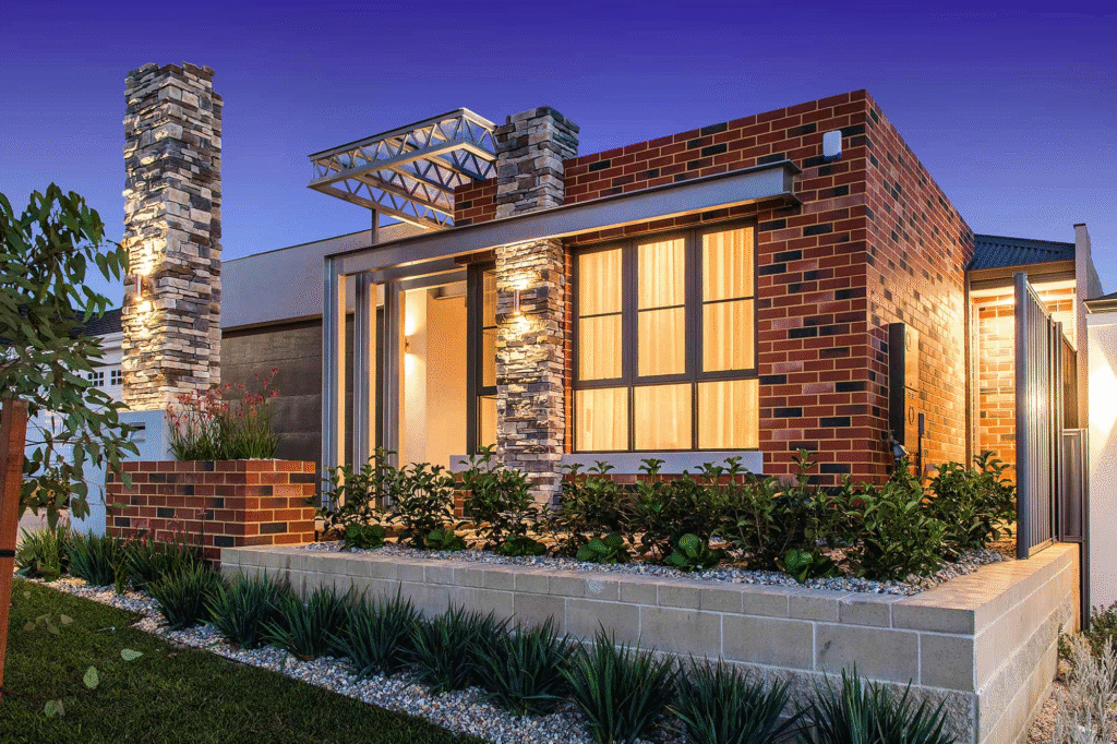 Modern single-storey brick house with large windows, illuminated stone pillars, and a neatly landscaped front garden by expert home builders, pictured at dusk under a deep blue sky.