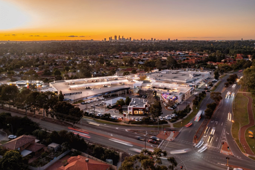 Aerial view of a shopping centre property with car parks at sunset, cars on surrounding roads, suburban houses, trees, and a city skyline in the distance—showcasing vibrant property in a thriving community.