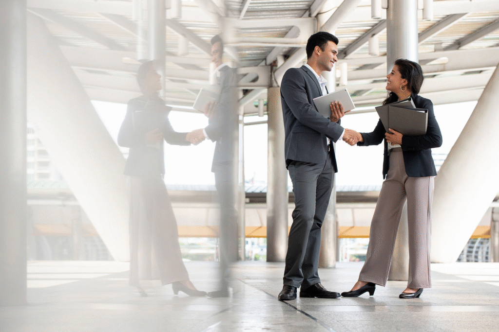 Two business professionals, a man and a woman, shake hands outdoors in a modern urban setting. Both are smiling, holding folders—highlighting their expertise in professional services—as their reflections shine in the nearby glass.