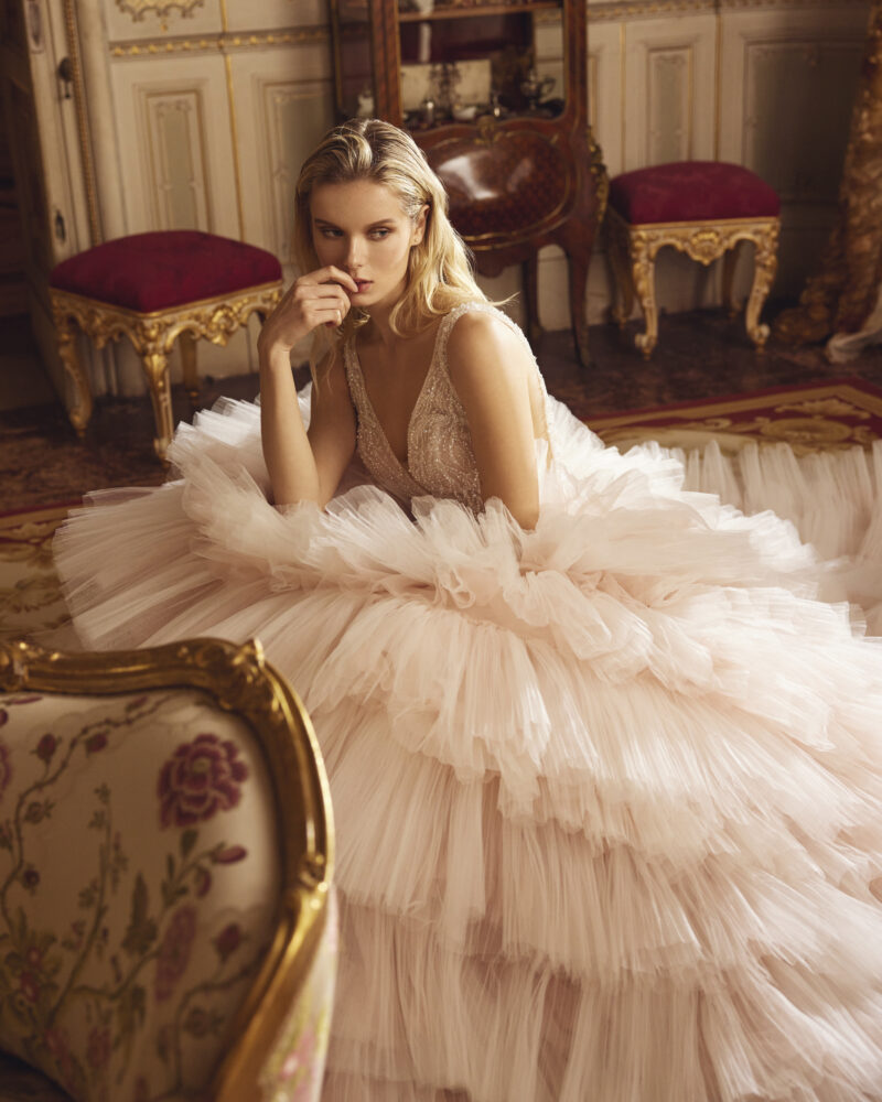A woman in a voluminous, ruffled blush pink gown embodies timeless fashion as she sits pensively in an ornate, vintage room with gold-trimmed furniture and rich red upholstery.