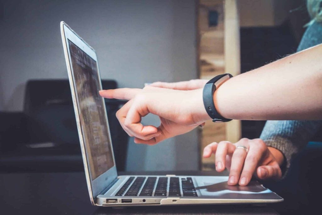 Two people interact with a laptop; one points at the screen while the other types on the keyboard, possibly discussing an SEO Perth strategy. Both hands are visible, one wearing a black wristband. The background is out of focus.