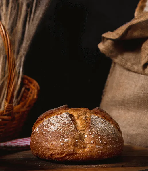 A round loaf of rustic Abhi’s Bread sits on a wooden surface. In the background, there’s a wicker basket with dried wheat and a hessian sack, all set against a dark backdrop.