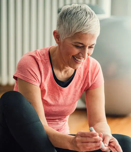 A smiling older woman with short grey hair wearing a pink sports shirt sits on the floor indoors, holding a medical device in her hand.