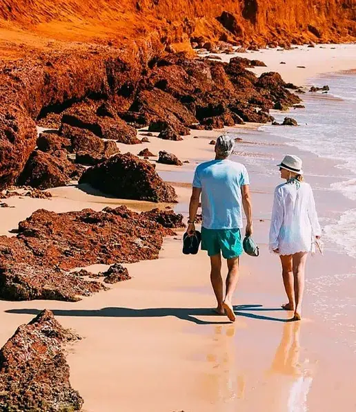 A man and a woman walk barefoot along a sandy beach with reddish cliffs and rocks, holding their shoes and hats, enjoying a sunny day by the sea—an experience like no other.