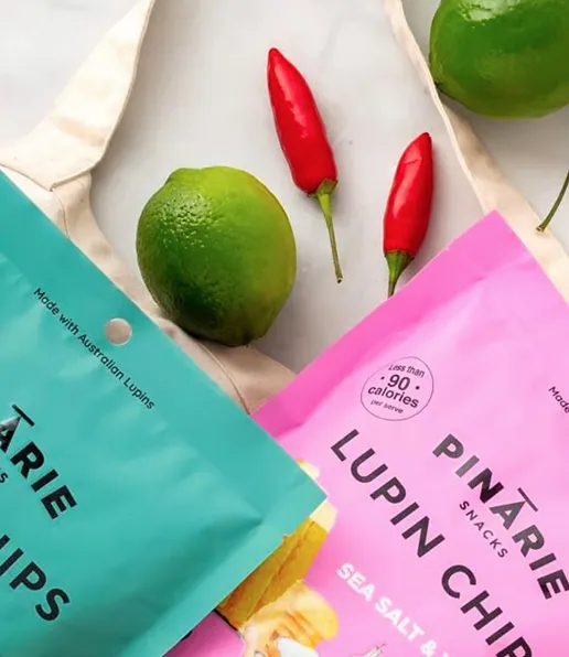 Two colourful crisp packets from Pinarie Foods, one teal and one pink, lie on a light surface next to a reusable tote bag, two red chilli peppers, and two whole limes.