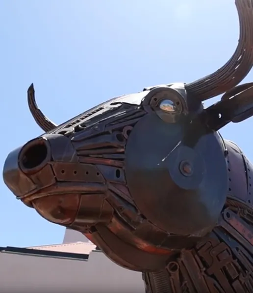 A close-up of a bull sculpture made from various pieces of scrap metal, including gears and pipes, stands outdoors under a clear blue sky in the Shire of Quairading.