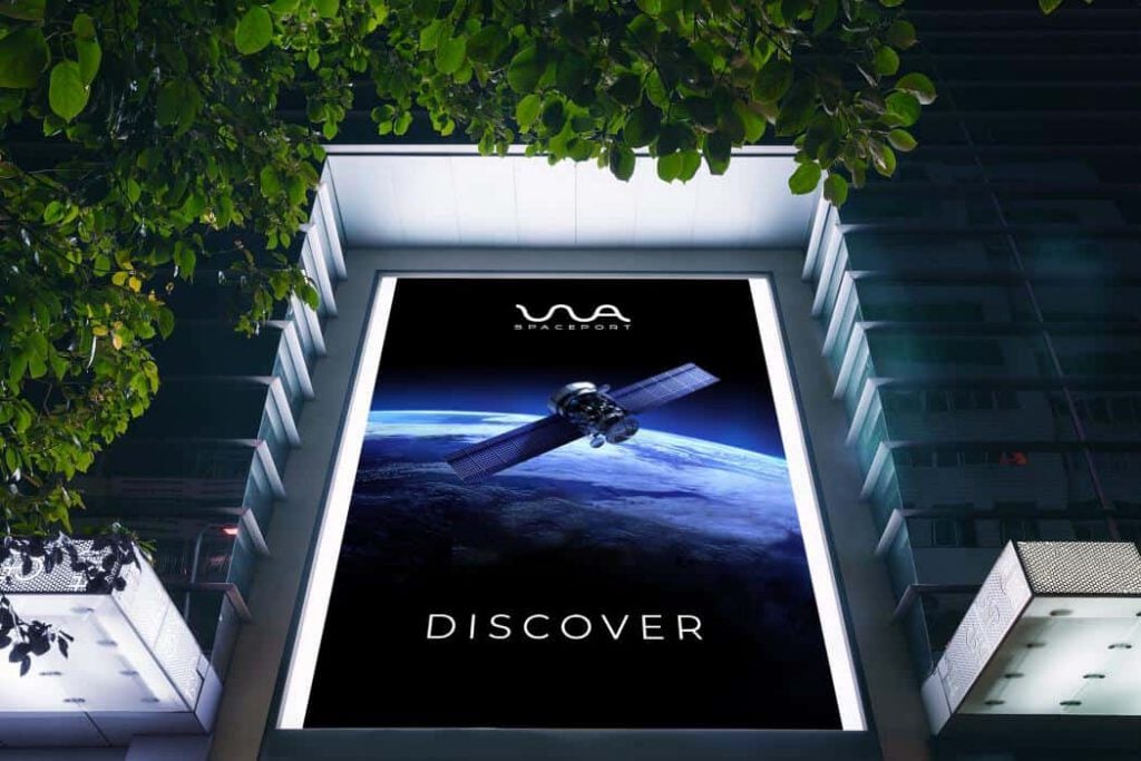A large illuminated hoarding on a city building displays an image of a satellite orbiting Earth with the word DISCOVER and the WA Spaceport logo, highlighting space launch Australia against a dark sky, framed by tree leaves above.