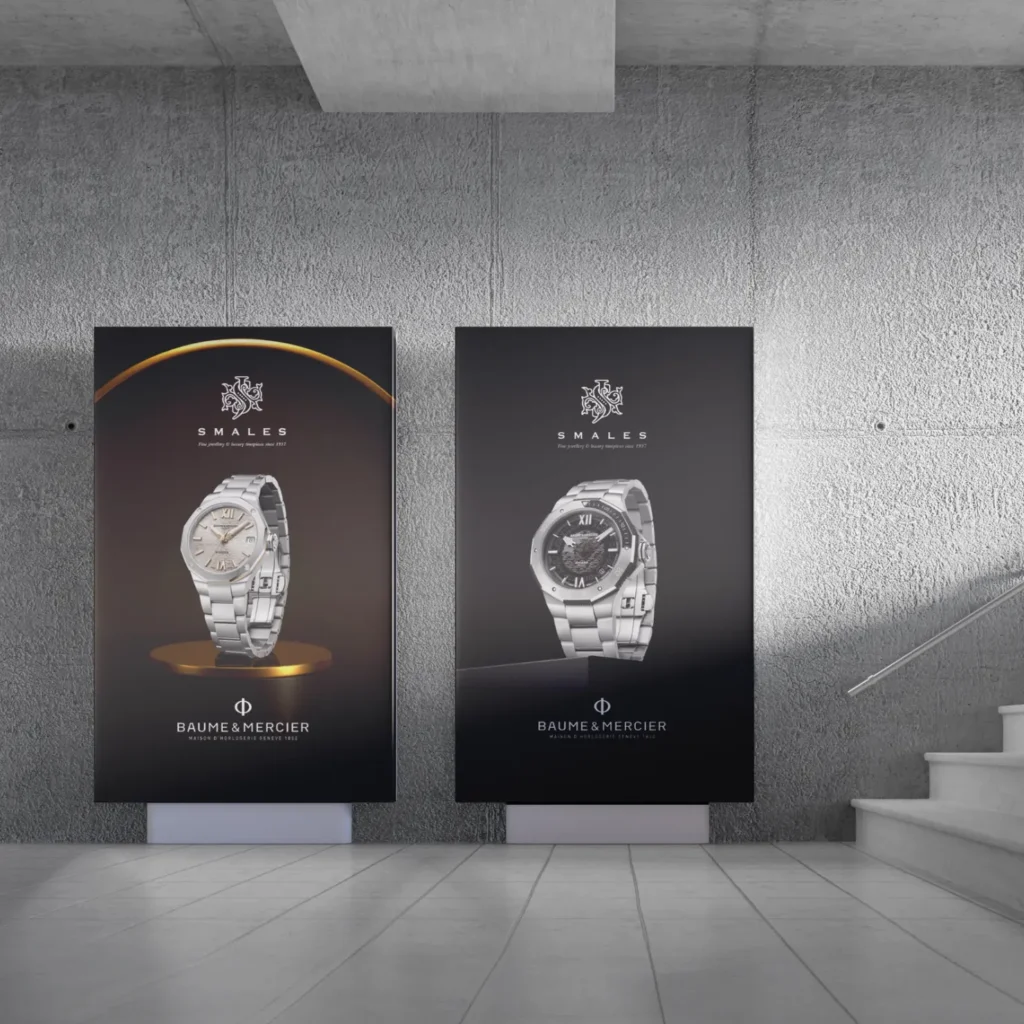 Two standing posters showcase luxury Baume & Mercier watches by Smales Jewellery, set against a modern, grey concrete wall with stairs visible on the right. Each poster features a different model from their luxury watches collection.