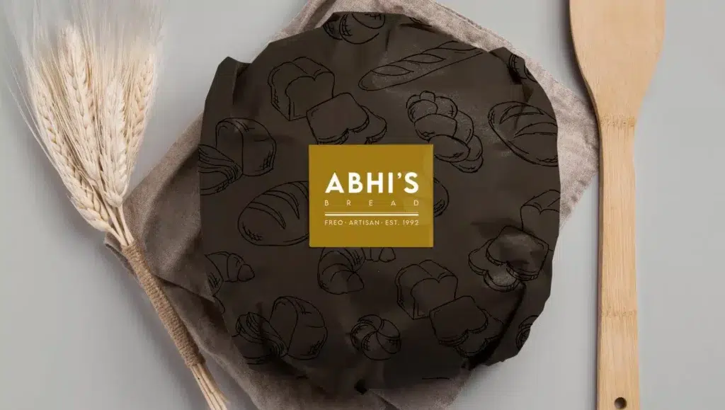 A round loaf covered with dark cloth printed with bread sketches, labelled Abhi’s Bread on a golden square. It sits on a beige linen, evoking the warmth of a bakery, with wheat stalks and a wooden spoon beside it.
