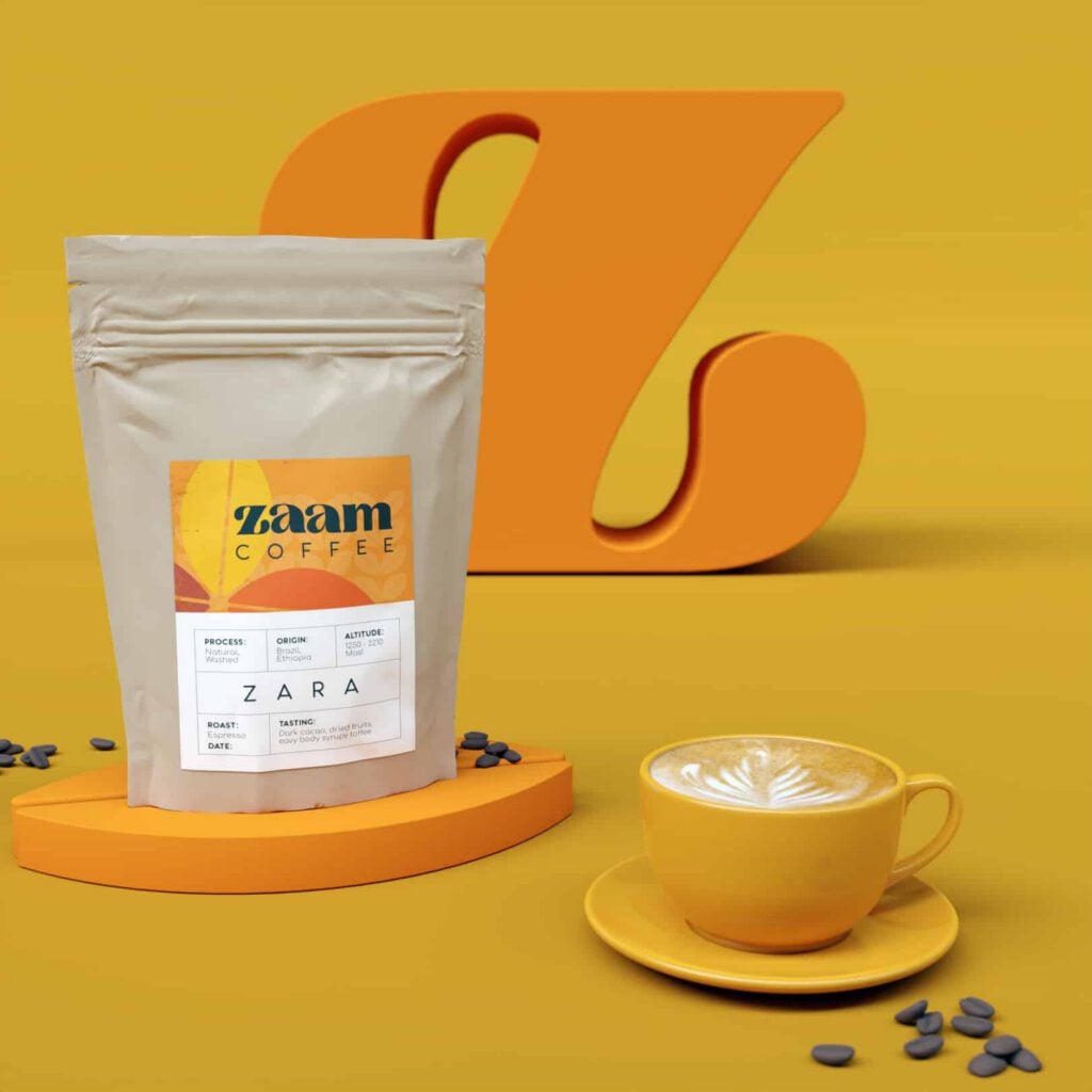 A bag of Zaam Coffee stands upright next to a yellow cup of latte with foam art, set against a vibrant yellow background with a large orange Z and coffee beans, highlighting the brand's dedication to expert coffee roasting.