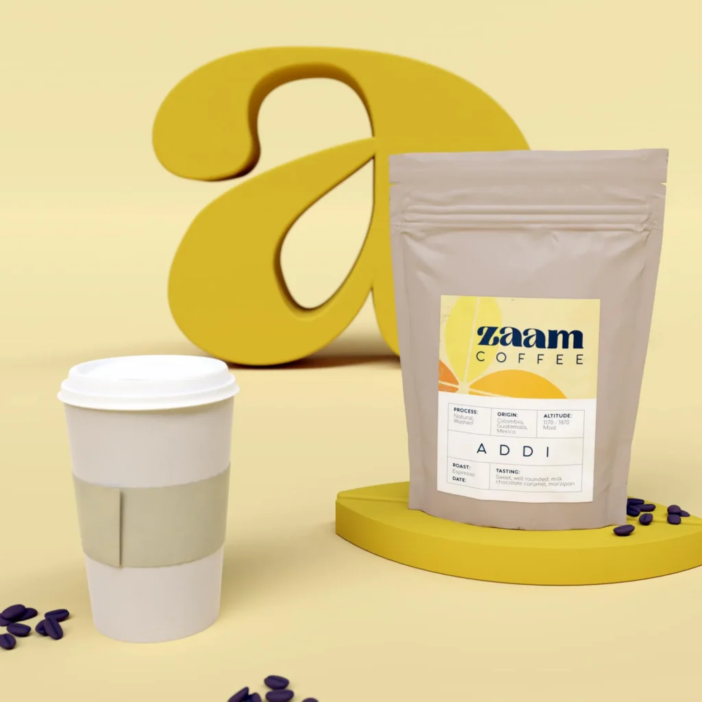 A takeaway coffee cup and a Zaam Coffee bag labelled ADDI are displayed in front of a large, yellow, stylised letter a with scattered coffee beans on a beige surface, capturing the essence of Zaam Coffee’s expert coffee roasting.