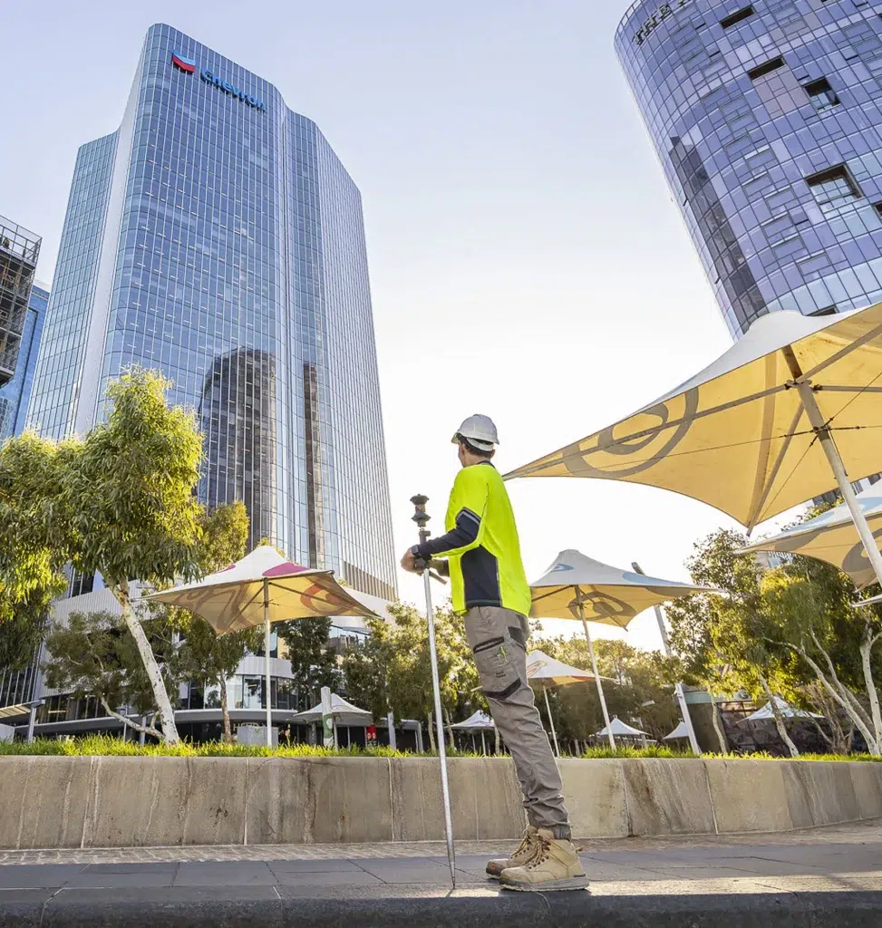 A construction worker in a high-visibility shirt and hard hat stands with Oracle Surveys equipment in an urban plaza, facing tall modern glass buildings and large outdoor parasols on a sunny day.