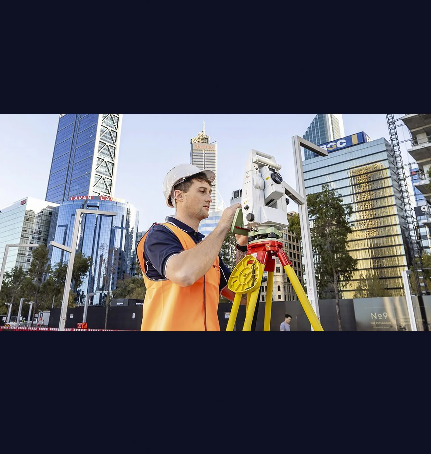 A construction worker in an orange vest and white helmet uses an Oracle Surveys instrument on a tripod in an urban area, surrounded by tall modern buildings.