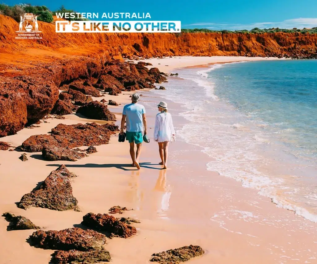 Two people walk barefoot along a sandy beach with orange-red cliffs and clear blue water in Western Australia. Text at the top reads, Western Australia—an adventure like no other.