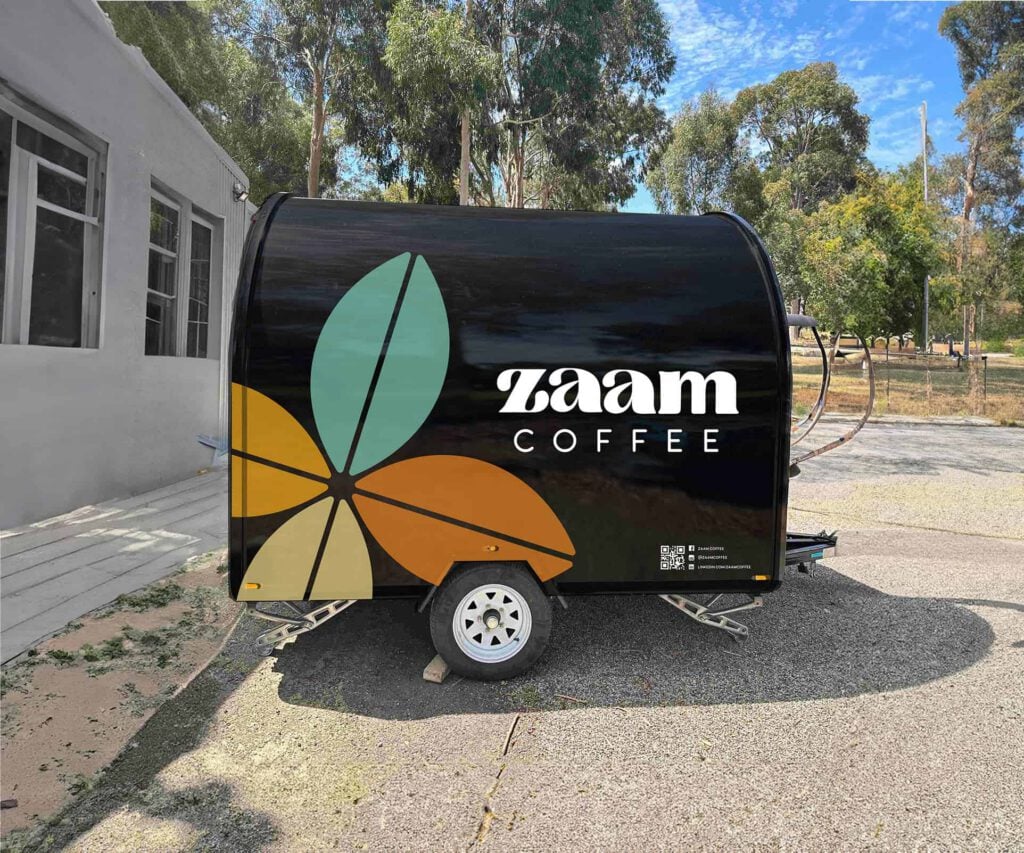 A black coffee trailer with large, colourful leaf graphics and the white text Zaam Coffee is parked outside a building near trees and a pavement on a sunny day, inviting passers-by to enjoy freshly roasted coffee beans.