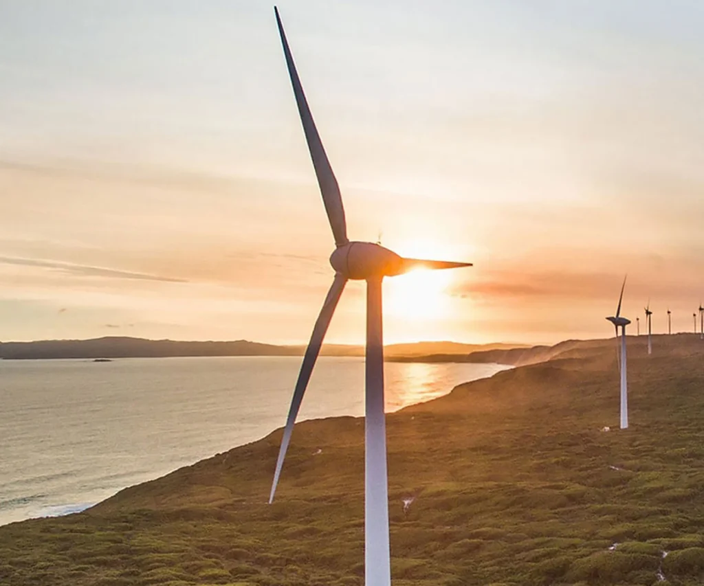 A wind turbine stands on a grassy coastal landscape at sunset, like no other view, with more turbines in the distance and the sea on the left under a colourful sky.