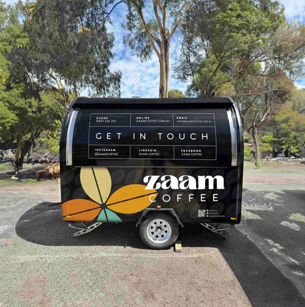 A black Zaam Coffee trailer with a colourful leaf graphic, coffee roasting details, and contact info is parked outdoors on a gravel area, surrounded by trees and greenery.