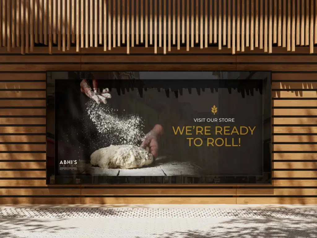 A bakery shopfront sign shows hands sprinkling flour over dough, with the text VISIT OUR SHOP. WE’RE READY TO ROLL! and Abhi’s Bread on a dark background, framed by wooden slats.