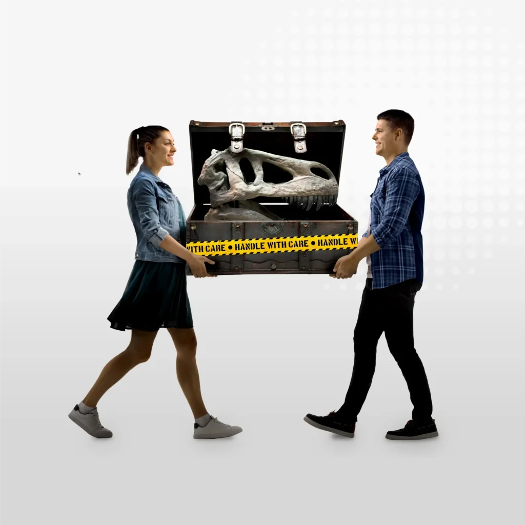 A smiling woman and man carry a large open case containing a dinosaur skull replica, wrapped with yellow HANDLE WITH CARE tape, as if ready for transport with Yello Lorry Hire, against a white background.
