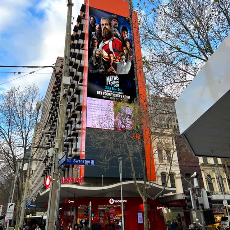 A tall, orange building on a city street displays a large vertical electronic billboard promoting Metro Comic Con, featuring images of actors in costume. People walk below; trees and street signs are visible.