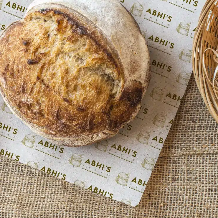 A round, rustic loaf of Abhi’s Bread sits on branded greaseproof paper with ABHIS printed repeatedly, next to a wicker basket on a hessian surface.