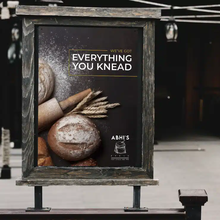A wooden-framed sign displays assorted artisan bread, wheat stalks, and rolling pins with the text WE'VE GOT EVERYTHING YOU KNEAD and Abhi’s Bread bakery on a dark background.