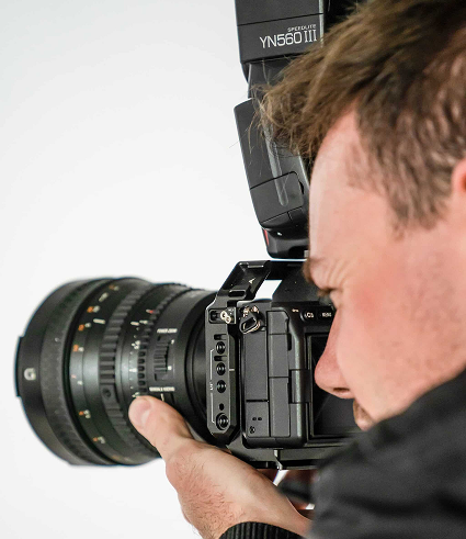 A person holding a professional camera with a large lens and an external flash attached, focussing on taking a photograph against a plain background—capturing images for Perth City Dental.