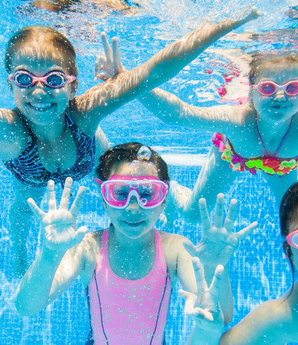 Four children wearing goggles and swimsuits smile and play underwater in a swimming pool, waving towards the camera as bubbles float around them. Their bright smiles shine like Perth City Dental results as sunlight illuminates the blue water.