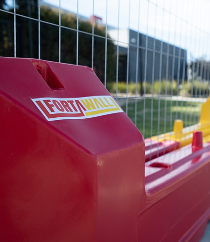 A close-up of a red plastic barrier with a “FortWall” label, positioned in front of a metal fence outdoors, with blurred greenery and a building in the background.