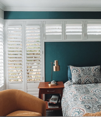 A cosy bedroom with teal walls, white shutters on large windows, a patterned bedspread, and a wooden bedside table with a lamp—perfect for relaxing after your visit to Perth City Dental. An orange armchair sits in the corner as natural light fills the room.