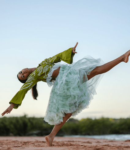 A woman in a green top and flowing skirt dances outdoors, balancing on one leg with arms extended gracefully, against a backdrop of sky and greenery—capturing the vibrant energy found in Perth, home to City Dental.