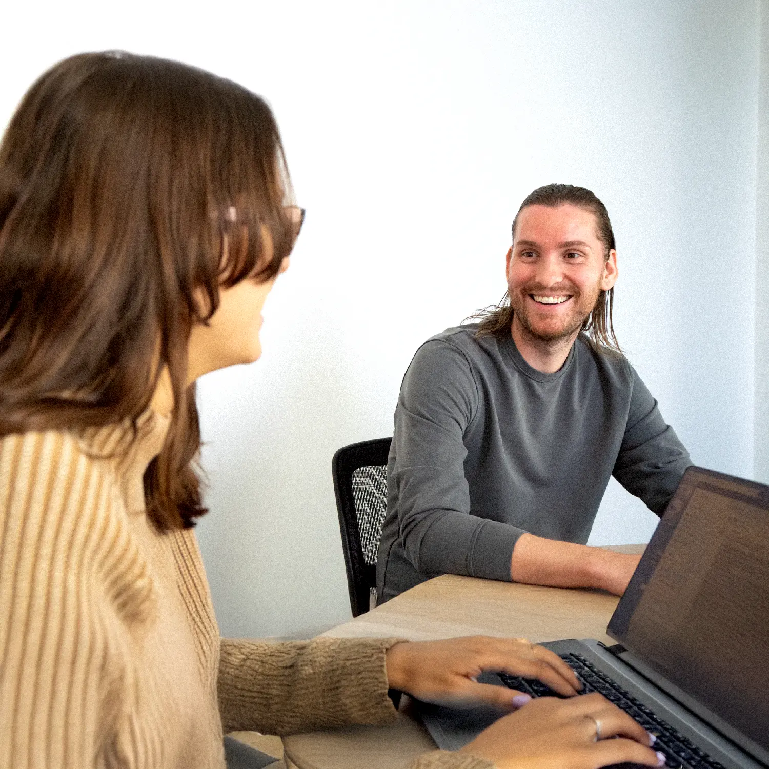 Two people sit at a table in a brightly lit office, smiling and talking. One types on a laptop as they discuss brand strategy, while the other looks on, engaged in the conversation. The background is plain and clean.