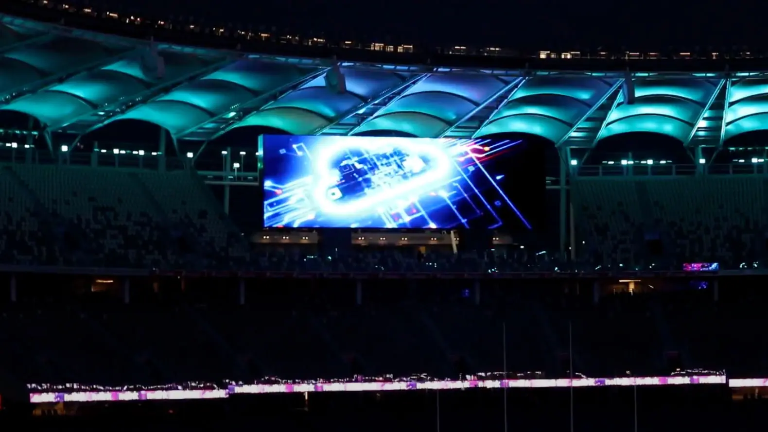 A large stadium at night with a brightly lit digital screen displaying blue and white graphics, under an illuminated teal roof. The seating areas are mostly dark, with some small visible crowds.