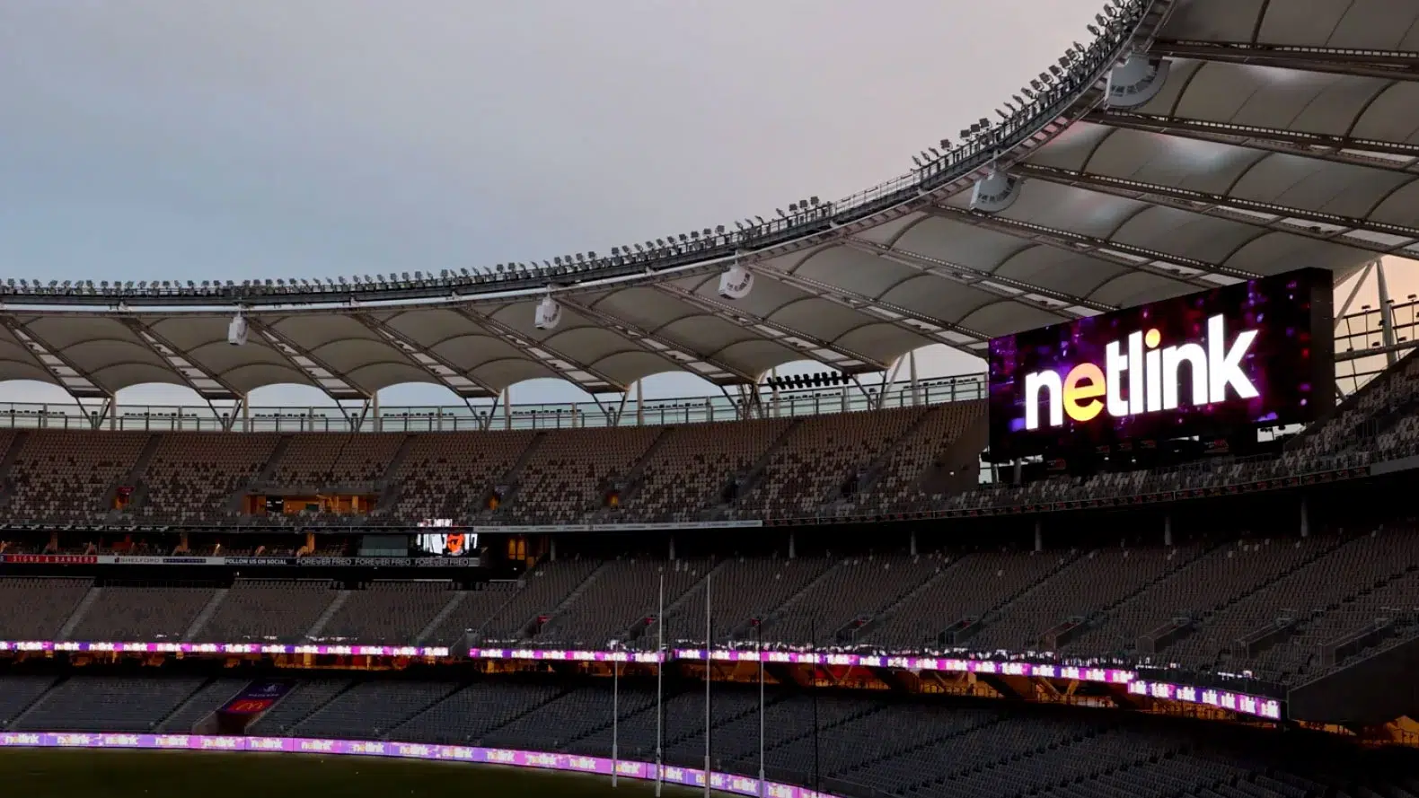 A large, mostly empty stadium with curved, white roof panels and tiered seating. A digital screen displays the word netlink in bright colours. The sky is overcast, and stadium lights line the roof.