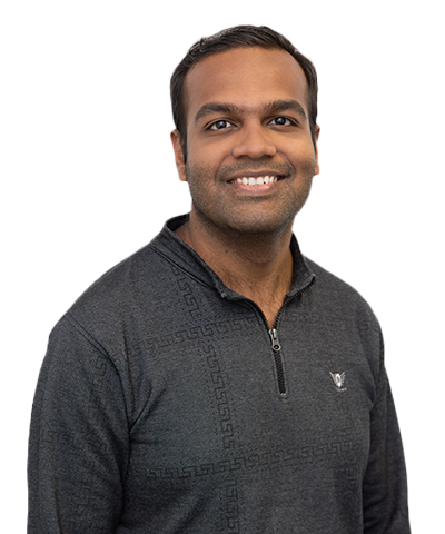 A man with short dark hair smiles at the camera. He is wearing a dark, patterned quarter-zip jumper and is posed against a plain, light background.