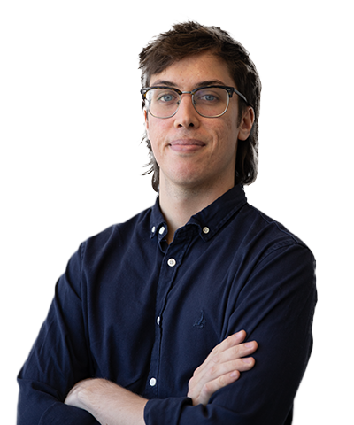 Harrison Sharrett, a young adult with glasses and brown hair, stands with arms folded in a dark blue button-up shirt, looking confidently at the camera against a plain white background.