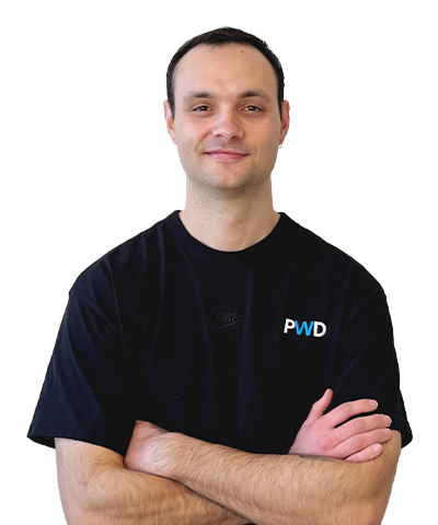 Tristram Vickers, a man with short dark hair, wearing a black T-shirt with PWD on it, stands with his arms folded and smiles slightly against a plain white background.