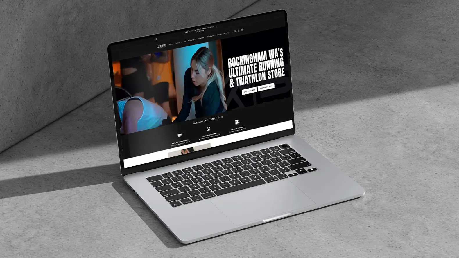 A silver laptop sitting on a grey surface displays the SBR Collective website, featuring a woman exercising and text that reads, Rockingham WA's Ultimate Running & Triathlon Store, with Shop Now and Find Out More buttons.
