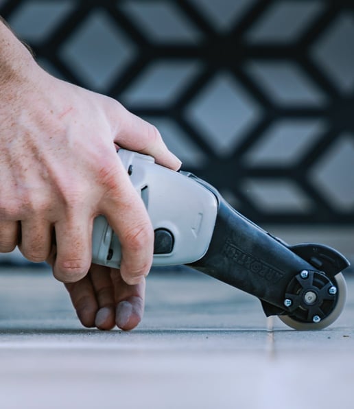 A hand holds and guides a rotary cutting tool along the edge of a tiled floor, making a precise cut—just the kind of detail AllSealed WA delivers. A geometric-patterned background is slightly blurred.