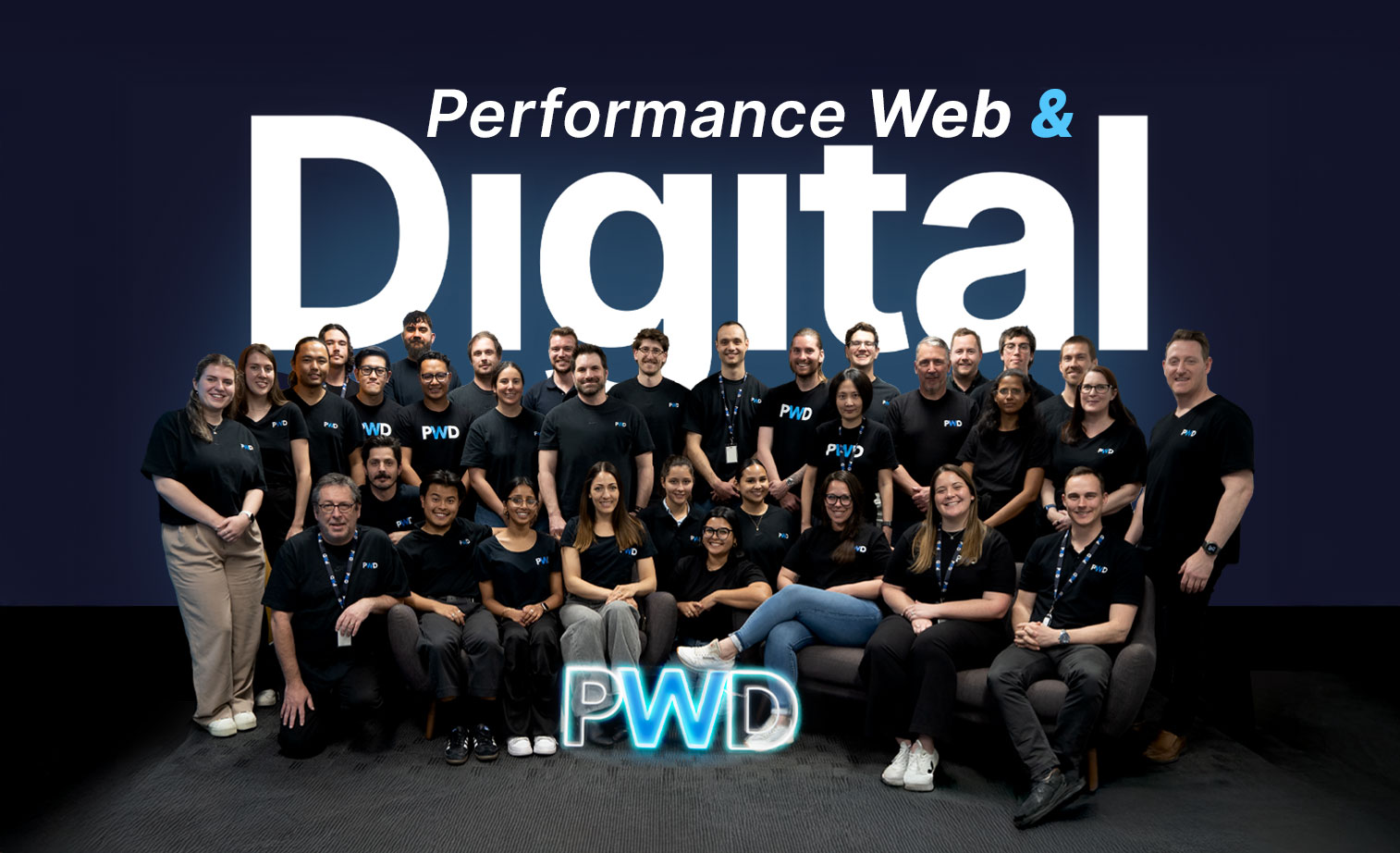 A group of people in matching black shirts pose and smile in front of a wall that reads Performance Web & Digital. A glowing PWD sign is displayed at the front.